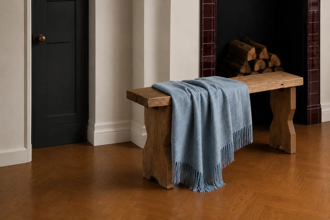 Light denim blue wool throw draped over a rustic wooden bench beside a fireplace in a warm contemporary interior.