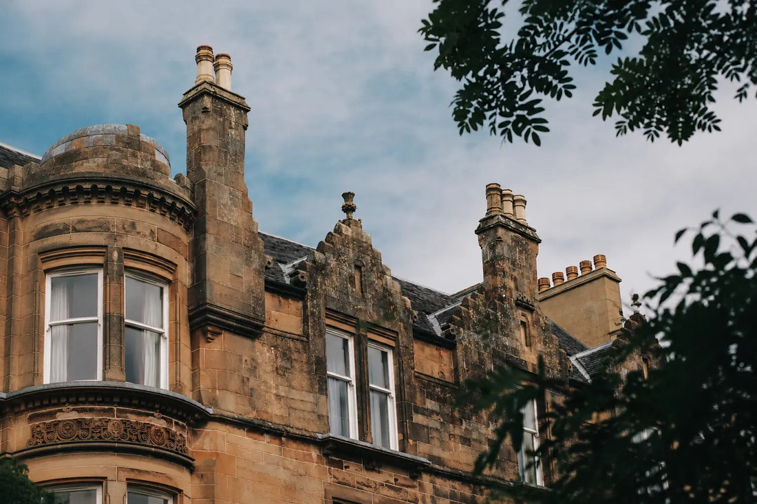 Historic Scottish sandstone architecture with ornate stonework, tall chimneys and traditional windows framed by leafy branches.