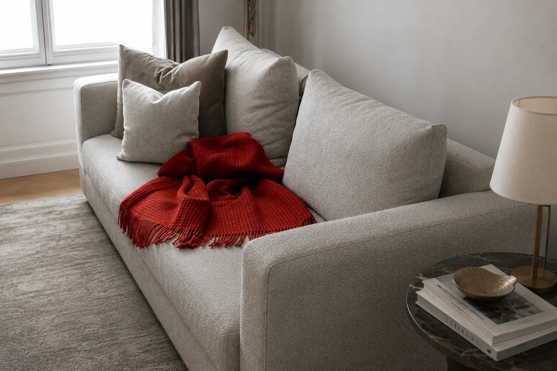 Copper red wool throw with fringed edges draped over a neutral textured sofa in a softly lit contemporary interior.