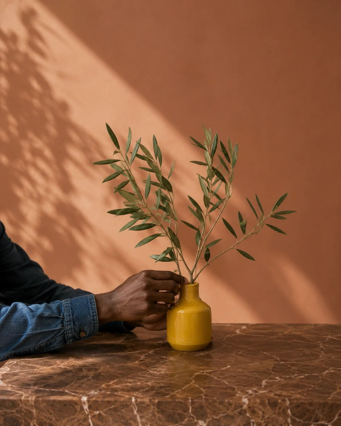 A small yellow ceramic vase holding green olive branches, placed on a brown marble table against a terracotta wall with soft leaf shadows.