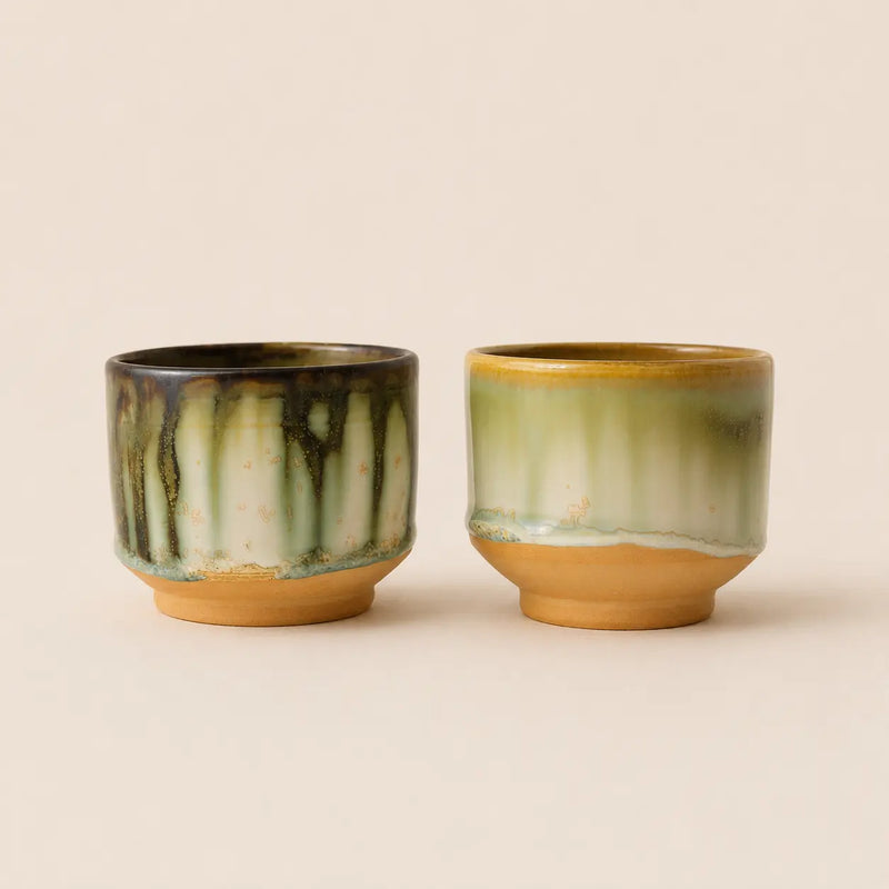 Two small glazed ceramic matcha bowls with a vertical drip design, one in green and black and the other in green and gold, set against a solid beige background.