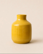 A small glossy ceramic vase in a warm mustard yellow color, centered against a solid, light beige background.