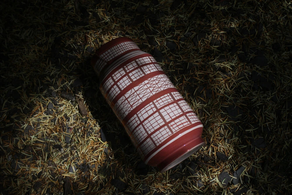 A cylindrical ceramic vase with copper and white geometric patterns lying on the ground among dry leaves and twigs.