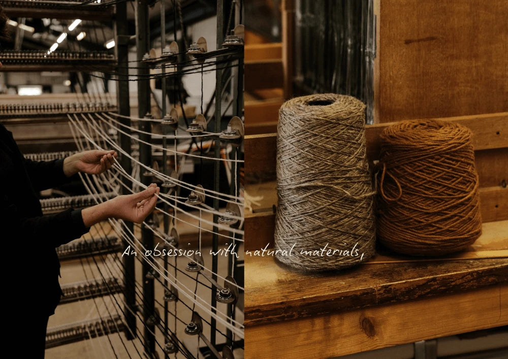 A person working with threads on a loom beside two large spools of natural yarn on a wooden surface.