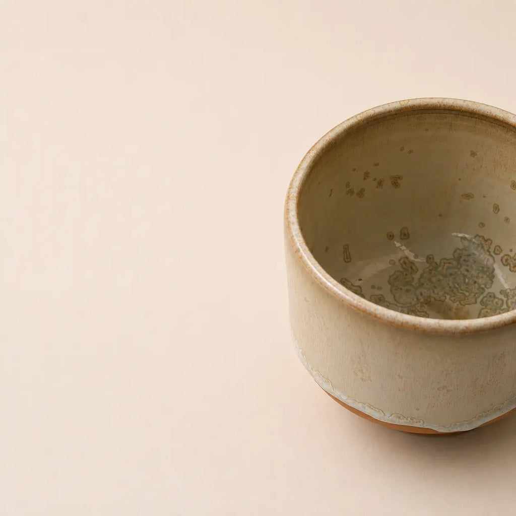 A handmade ceramic matcha bowl with a textured, speckled glaze in muted beige and brown tones, shown on a plain beige background with the cup positioned on the right side.
