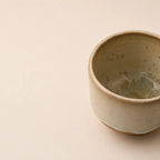 A handmade ceramic matcha bowl with a textured, speckled glaze in muted beige and brown tones, shown on a plain beige background with the cup positioned on the right side.