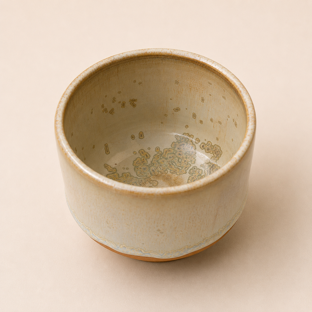 A handmade ceramic matcha bowl with a light beige and cream glaze, featuring subtle gold and brown speckled patterns inside and a rustic brown rim, set against a solid beige background.