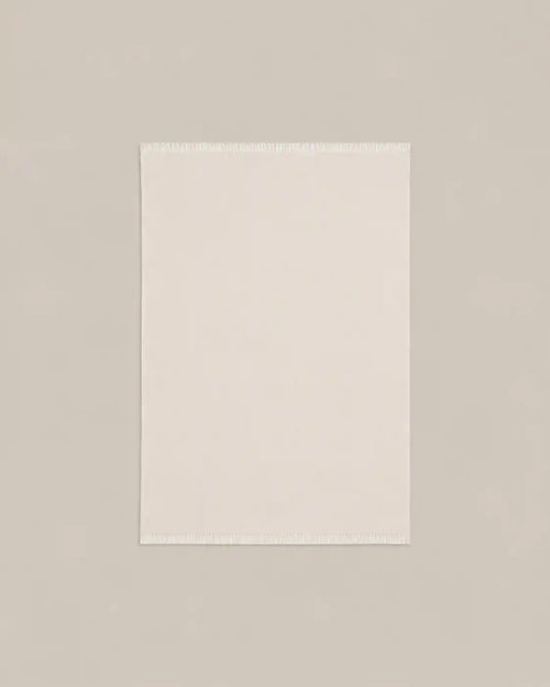 A plain cream-coloured cashmere blanket throw with fringed edges, shown flat against a gray background.