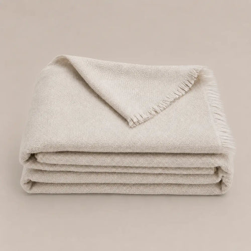 A folded cream-colored woven throw blanket with fringed edges, displayed on a solid, warm beige background.