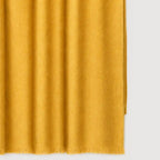 A close-up of a folded saffron yellow cashmere blanket, soft vertical folds and short fringe along the bottom edge, shown against a light gray background.