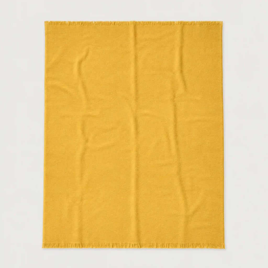 A rectangular mustard yellow throw blanket with fringed edges, laid flat against a light gray background.