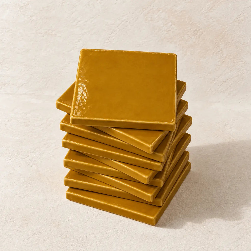 A stack of square ceramic tile in a glossy amber light brown finish, set against a textured off-white background.