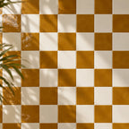 A checkered pattern of glossy amber light brown and off-white square tiles, with soft leaf shadows cast across the surface.