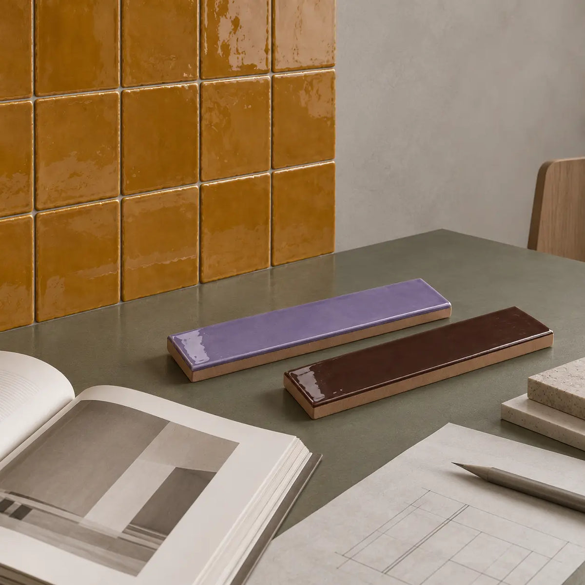Two glossy rectangular ceramic tile samples, one lavender purple and one dark brown, displayed on a gray tabletop next to an open book and architectural sketches, with a wall of mustard yellow tiles in the background.