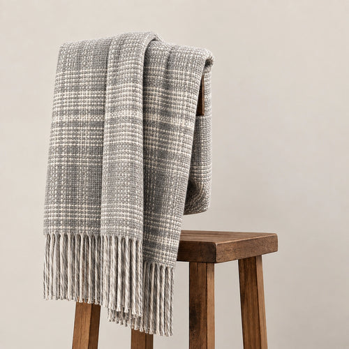 A gray and off-white woven throw blanket with fringe, draped over a wooden stool against a plain beige wall.