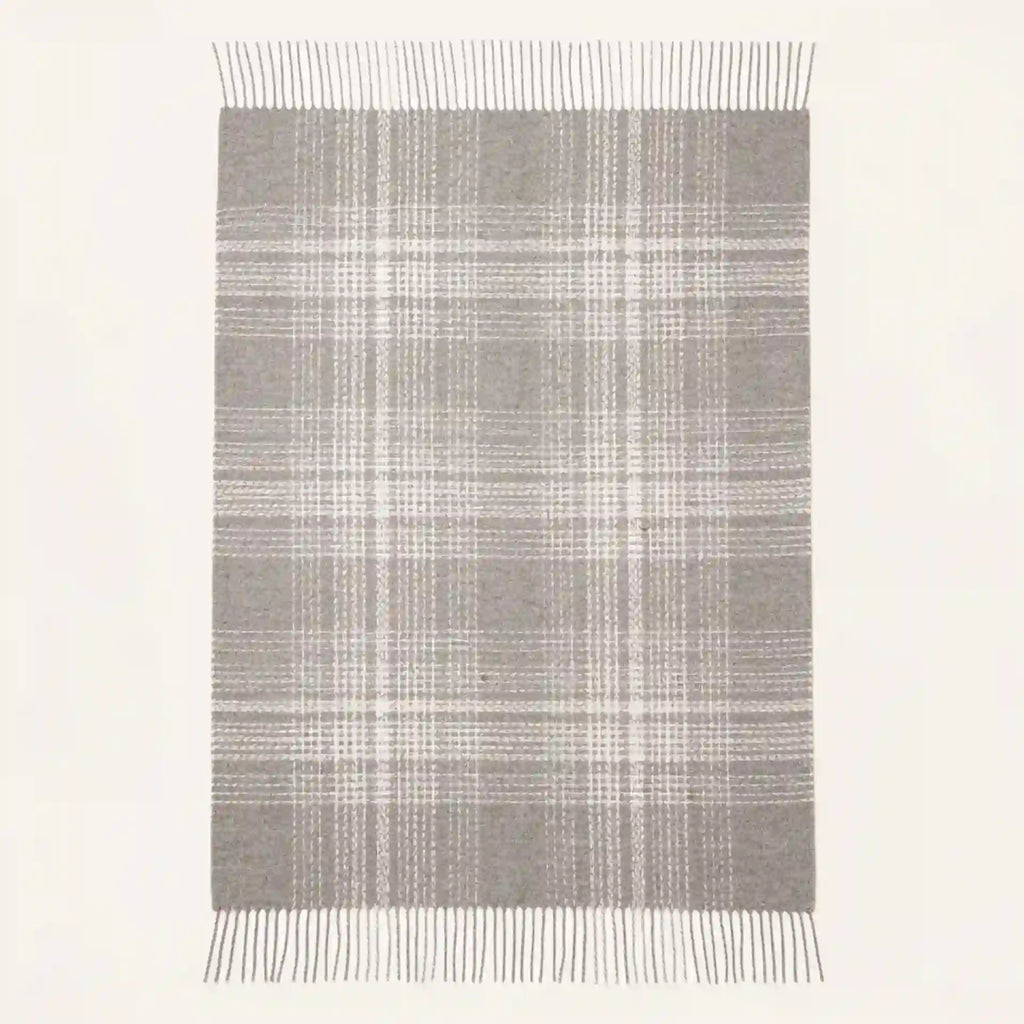 A rectangular throw blanket with a gray and white plaid pattern and fringe on the top and bottom edges, shown against a plain off-white background.