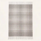 A rectangular throw blanket with a gray and white plaid pattern and fringe on the top and bottom edges, shown against a plain off-white background.