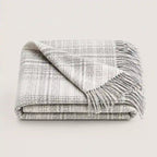 A folded woven throw blanket with fringe, shown on a plain light background. The blanket has a textured plaid pattern in off-white and gray.