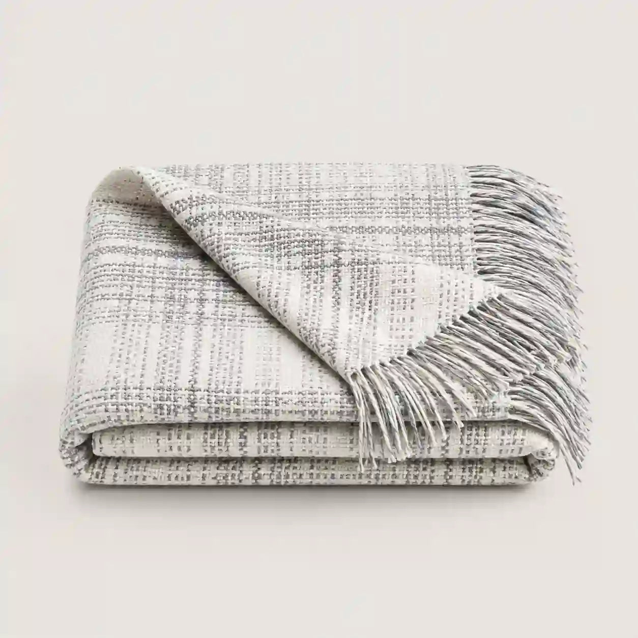 A folded woven throw blanket with fringe, shown on a plain light background. The blanket has a textured plaid pattern in off-white and gray.