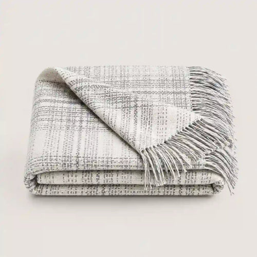 A folded woven throw blanket with fringe, shown on a plain light background. The blanket has a textured plaid pattern in off-white and gray.