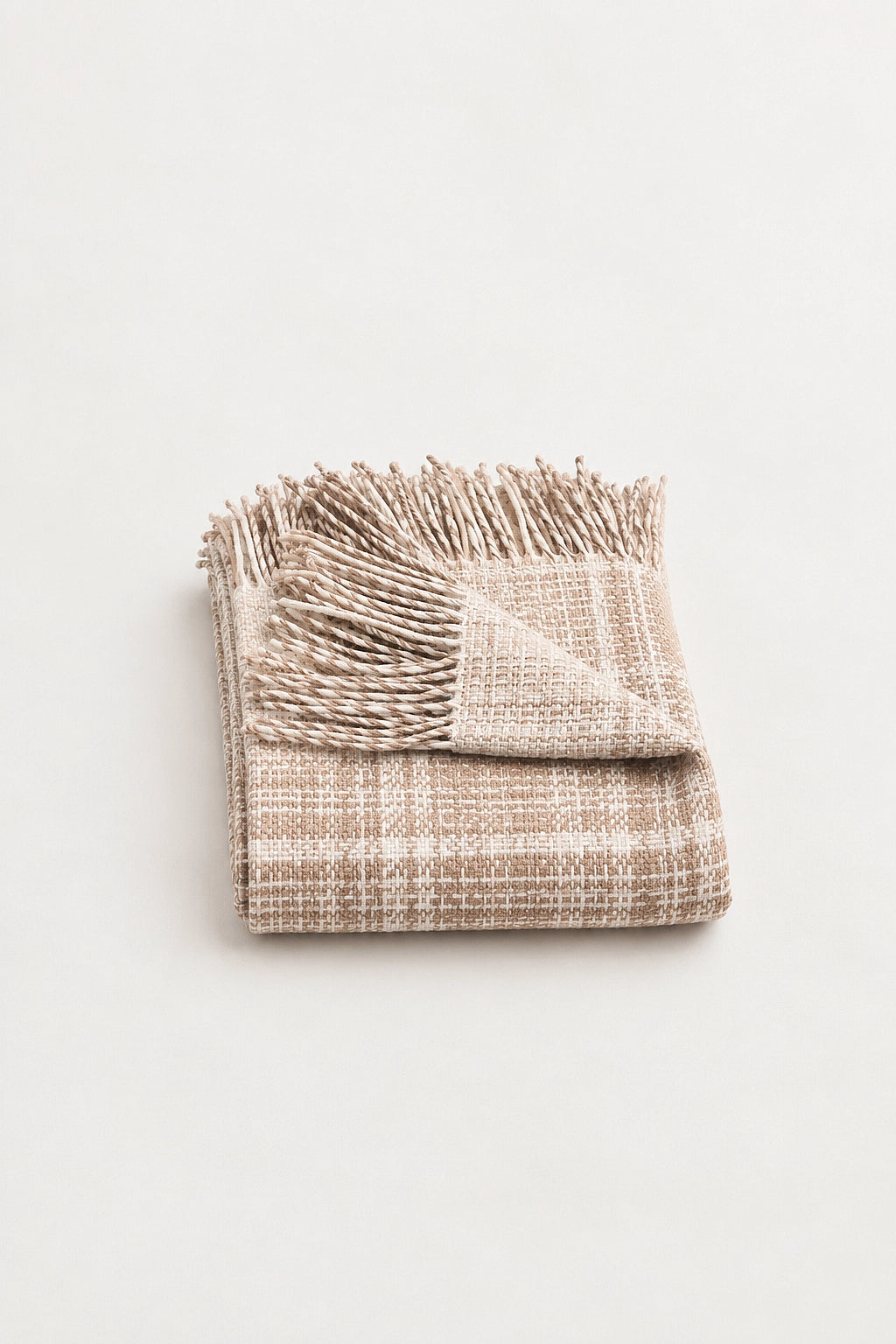 A folded woven throw blanket with fringed edges, featuring a soft beige and light brown plaid pattern, photographed on a clean off-white background.
