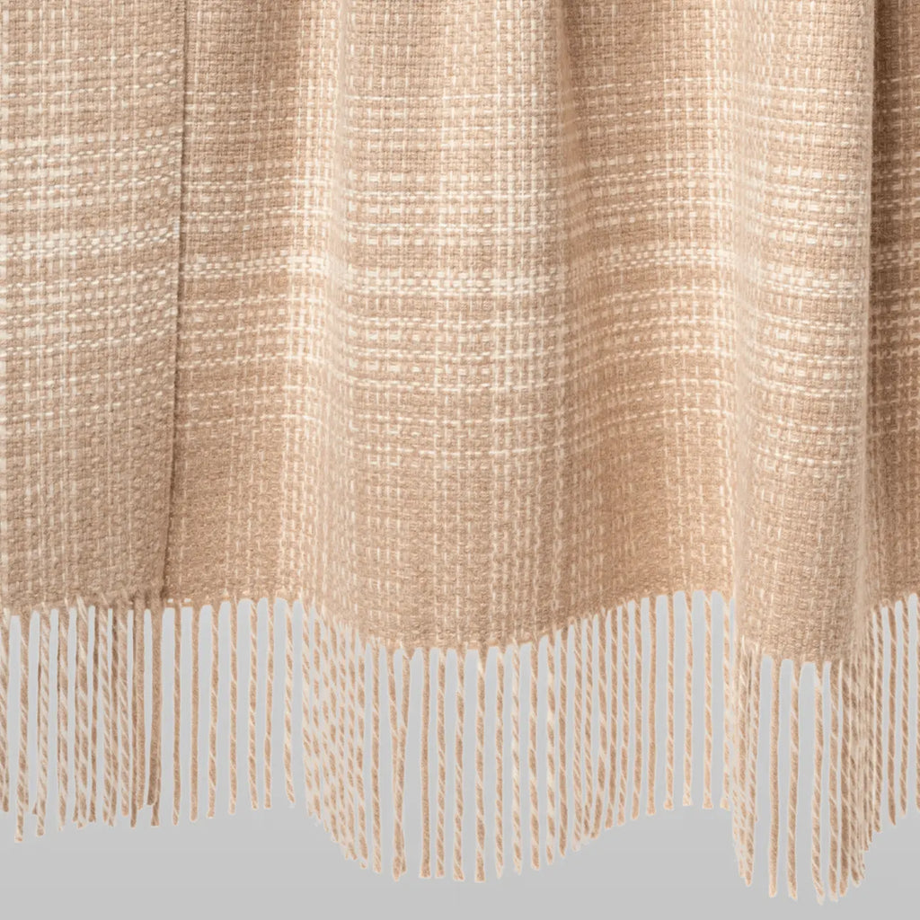 A close-up of a woven throw blanket in a warm beige and cream plaid pattern with a fringed hem, shown against a light gray background.