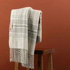 A cream and gray woven throw blanket with fringe, draped over a dark wooden stool against a warm terracotta background.