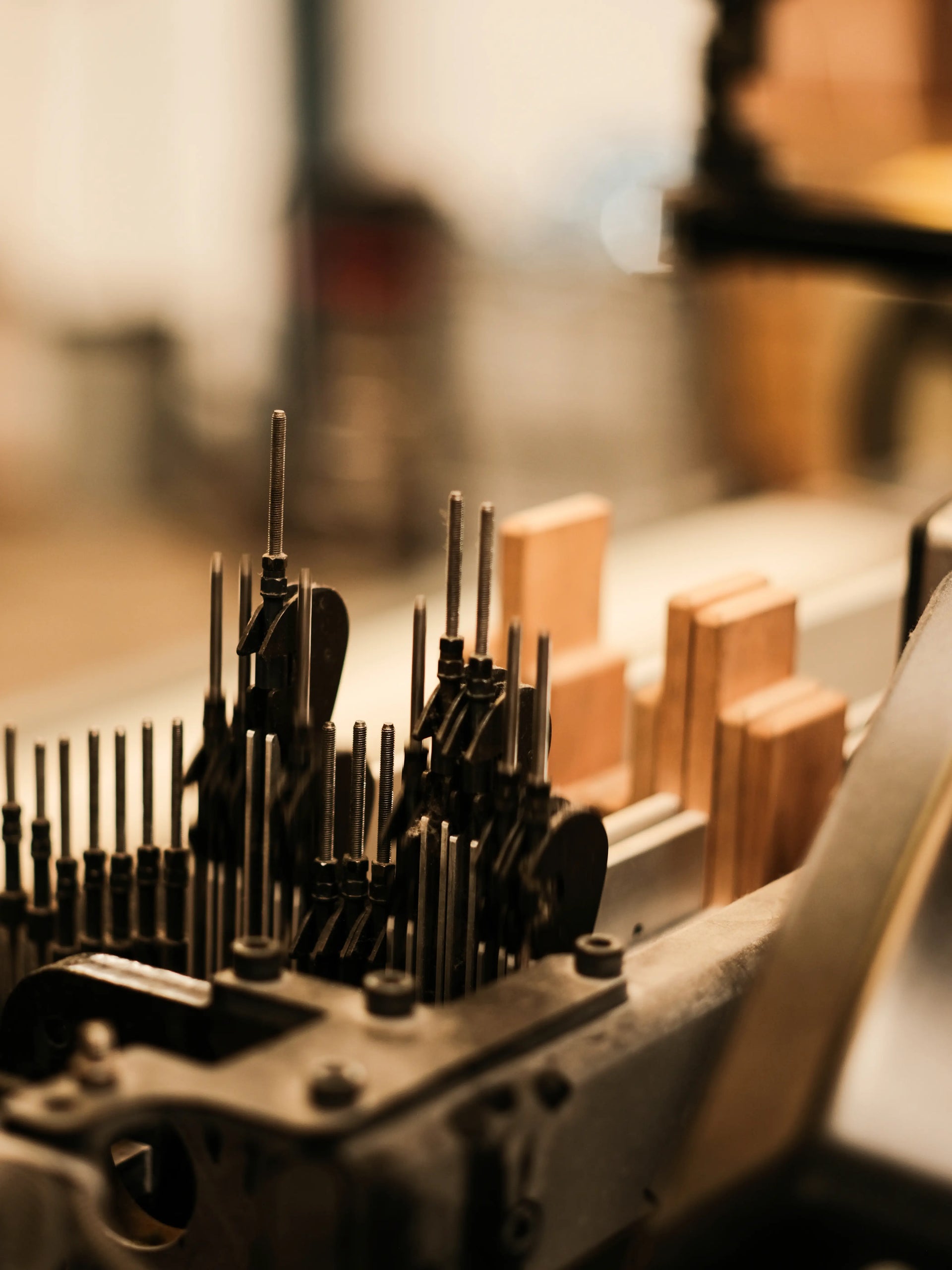 A close-up, shallow depth-of-field view of a machine holding numerous thin metal rods or pins, with small wooden blocks in the background, shot in warm, golden-brown tones.