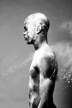 Black and white artistic photograph of a man in profile covered in white powder or snow, with spray particles visible against a dark gray background.