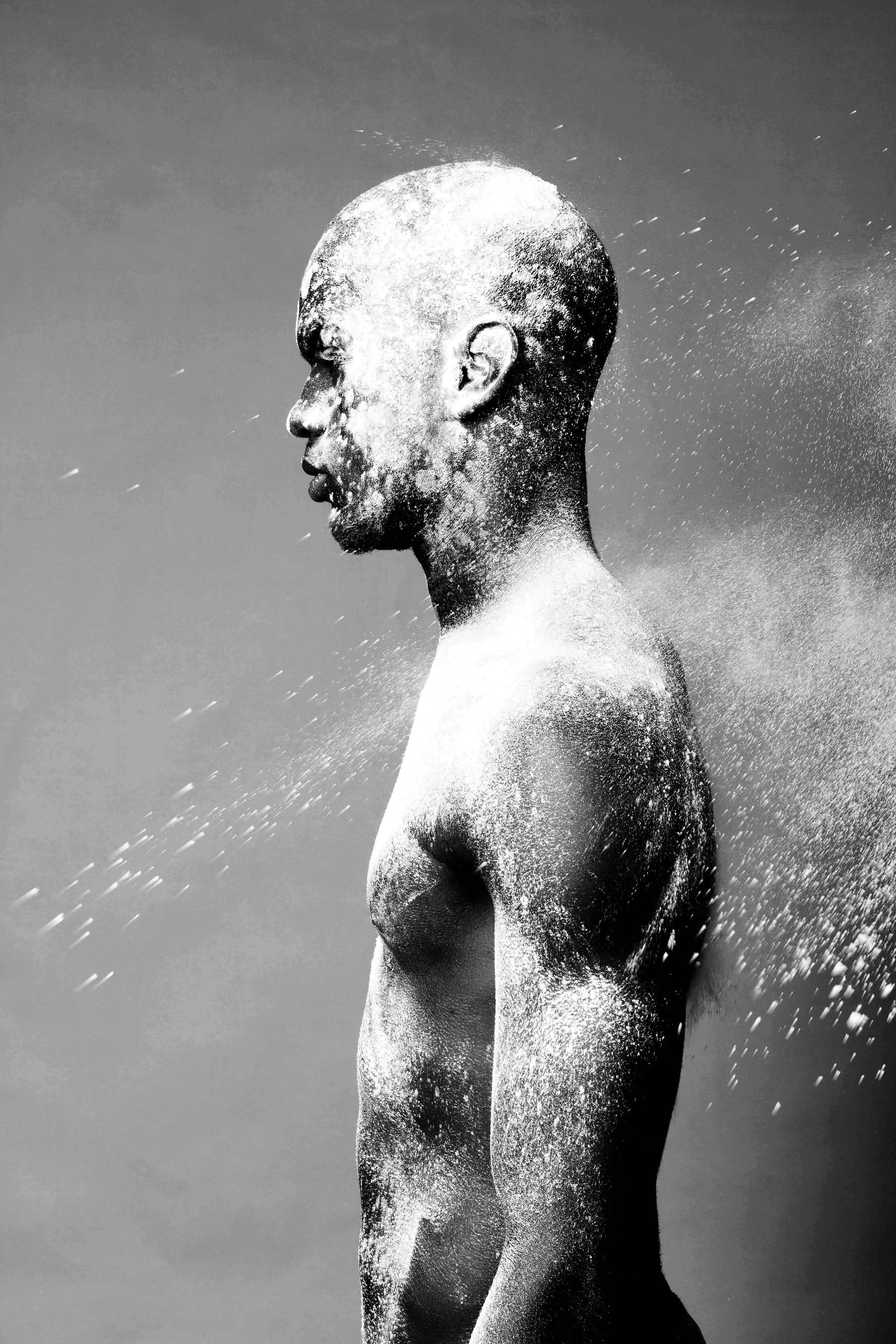 Black and white artistic photograph of a man in profile covered in white powder or snow, with spray particles visible against a dark gray background.