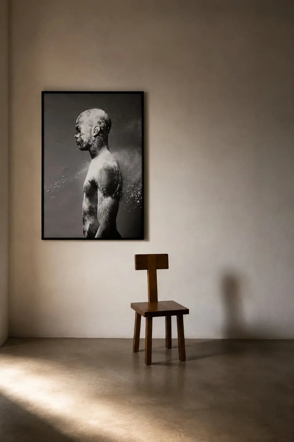 A framed black and white art print of a classical sculpture bust hangs on a textured beige wall above a minimalist wooden chair in a sunlit room.