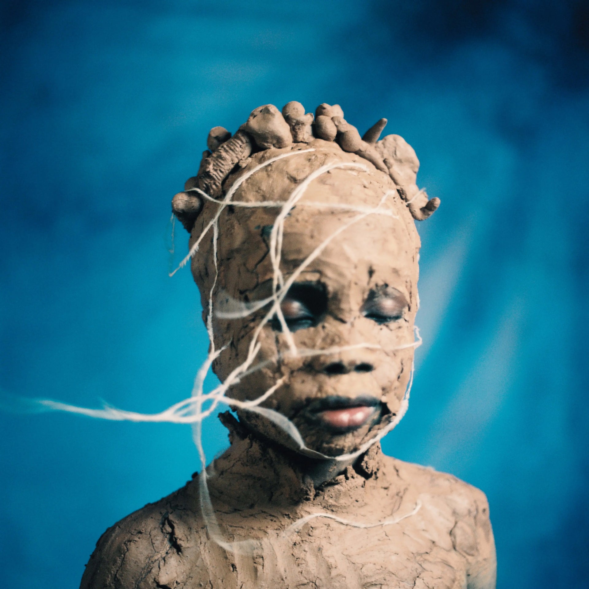 A sculptural clay bust of a face with eyes and lips, wrapped in white string against a vivid blue background.