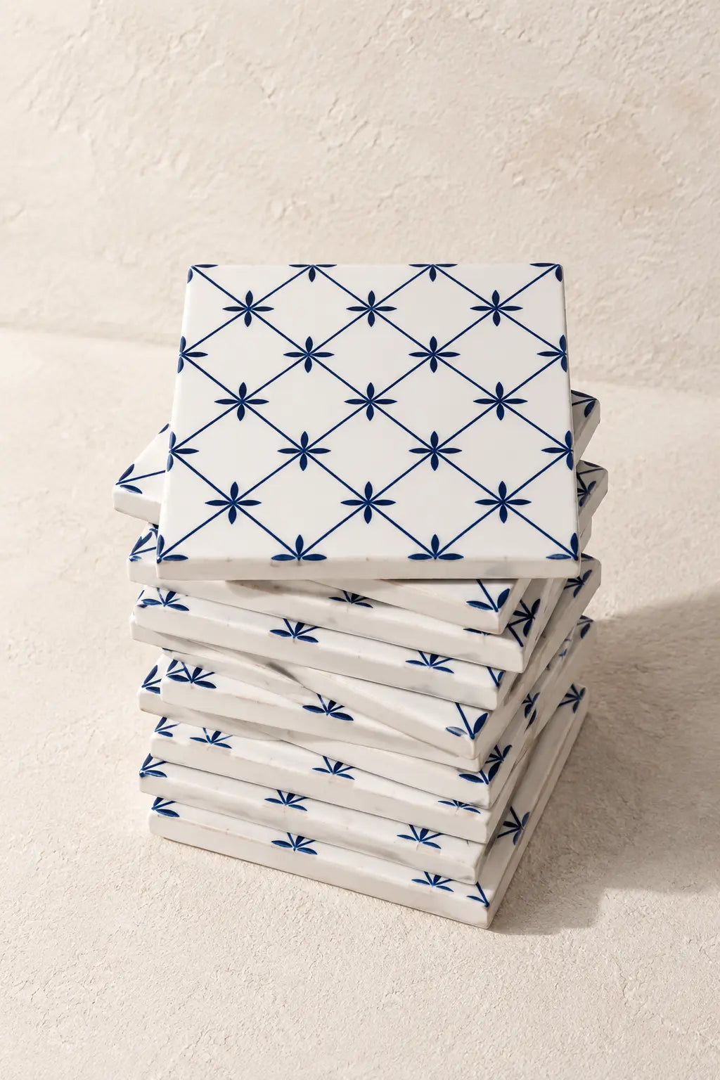 A stack of square white coasters with a blue geometric starburst pattern, photographed on a textured beige surface.