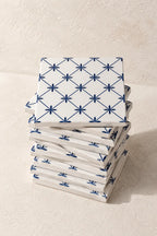 A stack of square white coasters with a blue geometric starburst pattern, photographed on a textured beige surface.