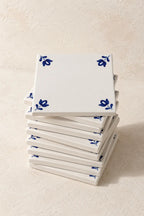 A stack of square white ceramic coasters with a blue floral pattern in the corners, set against a textured beige background.