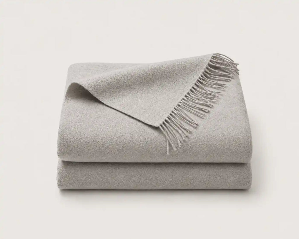 A folded light gray throw blanket with fringed edges, stacked on a plain off-white background.