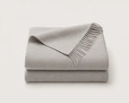 A folded light gray throw blanket with fringed edges, stacked on a plain off-white background.