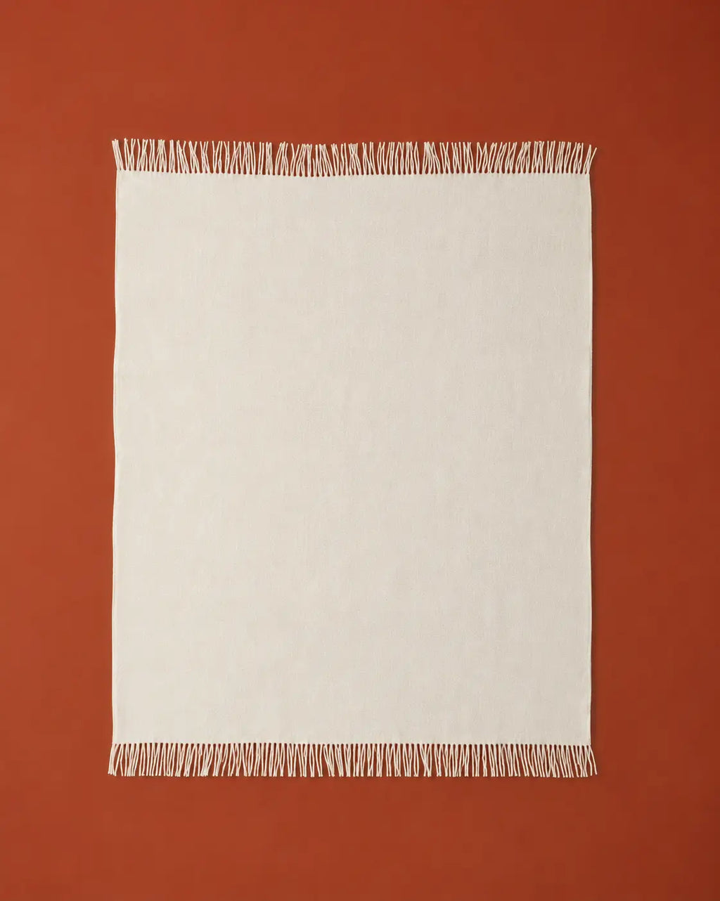 A rectangle off-white throw blanket with fringed edges, laid flat against a solid terracotta background.