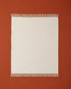 A rectangle off-white throw blanket with fringed edges, laid flat against a solid terracotta background.
