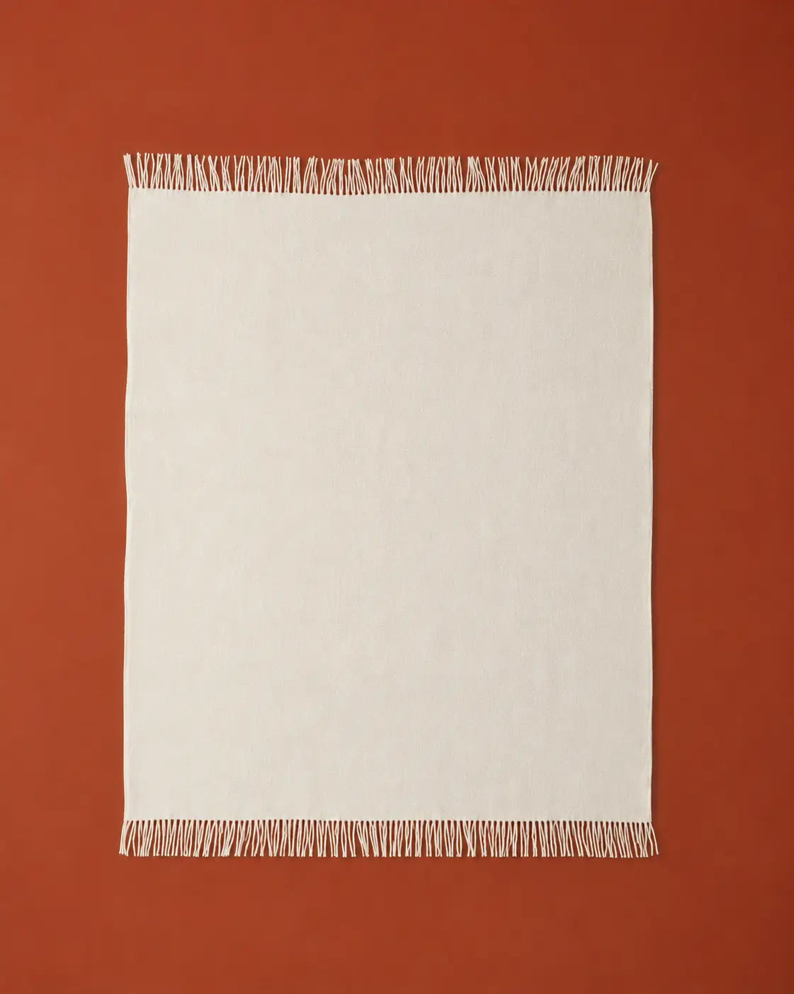 A rectangle off-white throw blanket with fringed edges, laid flat against a solid terracotta background.
