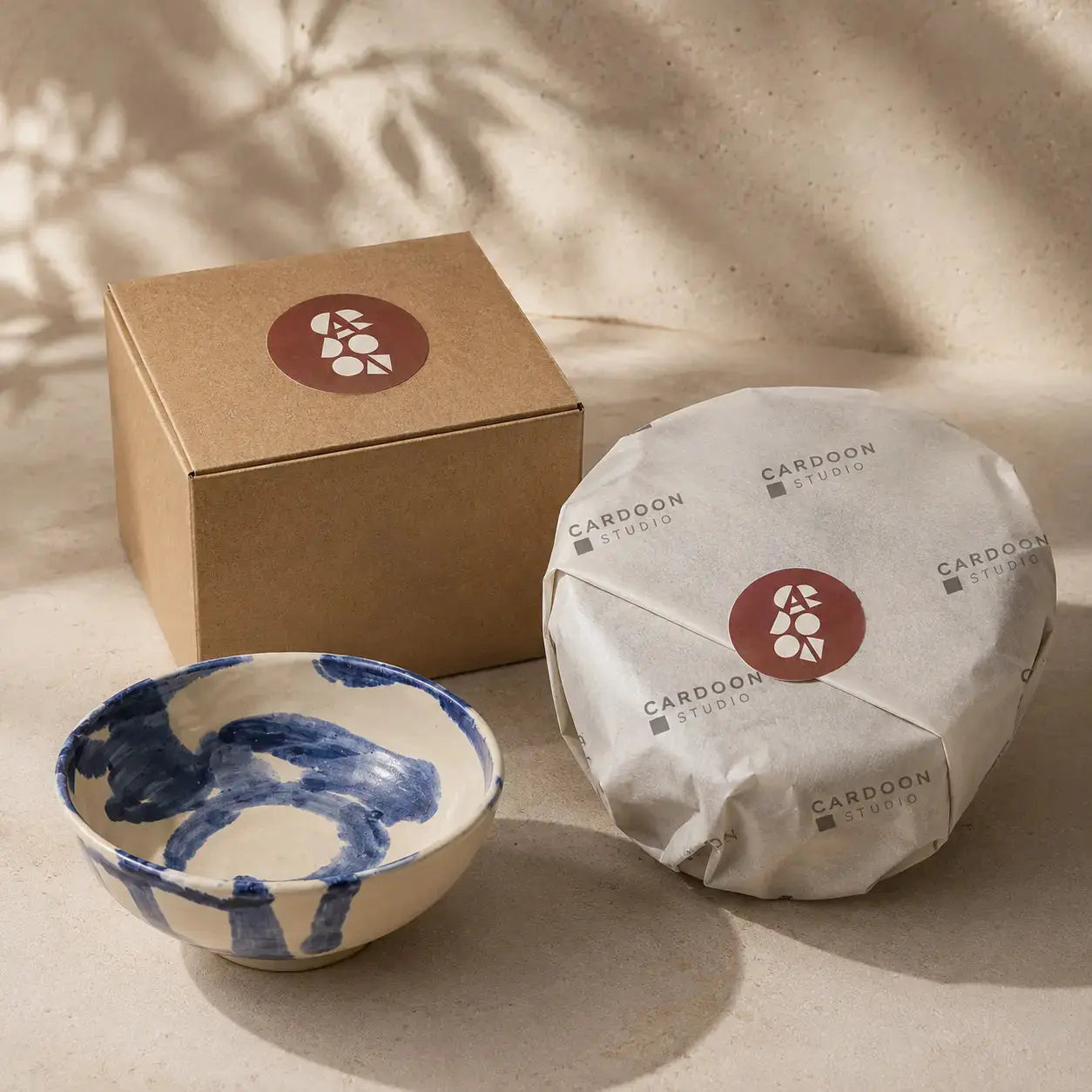 Ceramic bowl with blue patterns, wrapped product, and cardboard box on a neutral background