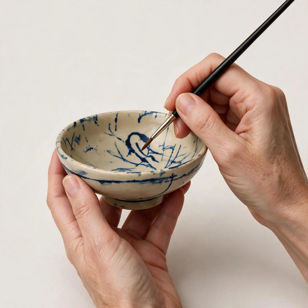 A ceramic bowl with a beige base and blue hand-painted brushstroke pattern, held by two hands while a third hand uses a fine paintbrush to add more detail inside the bowl, against a plain off-white background.