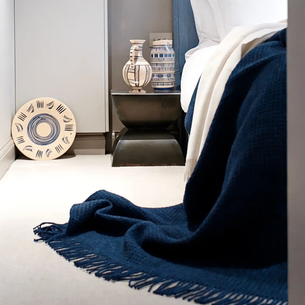 A navy blue textured throw blanket with fringe is draped over the corner of a bed in a modern bedroom setting.