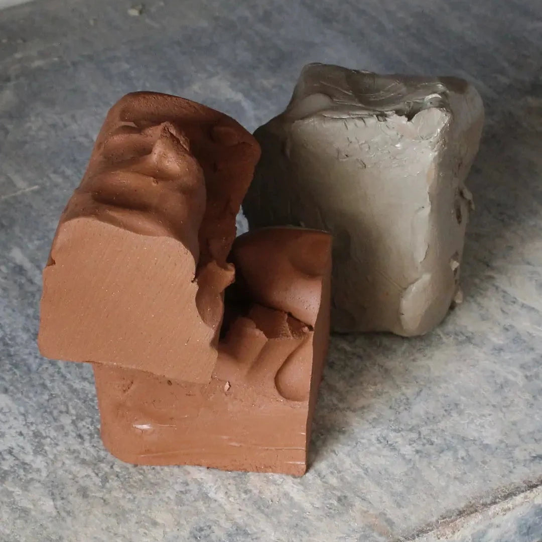 Two rough blocks of clay or soap, one terracotta brown and one light gray, resting on a dusty gray work surface.