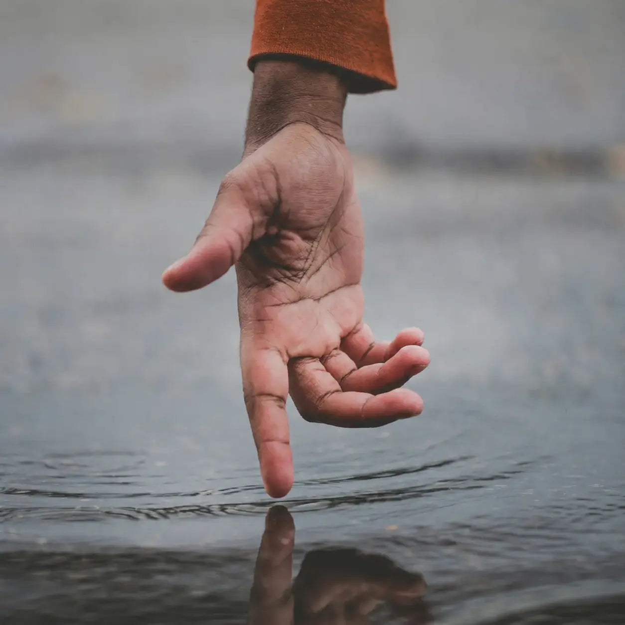 A close-up of a human hand reaching toward water, with the wrist showing a rust-orange sleeve, set against a blurred gray background and a faint reflection on the water surface.