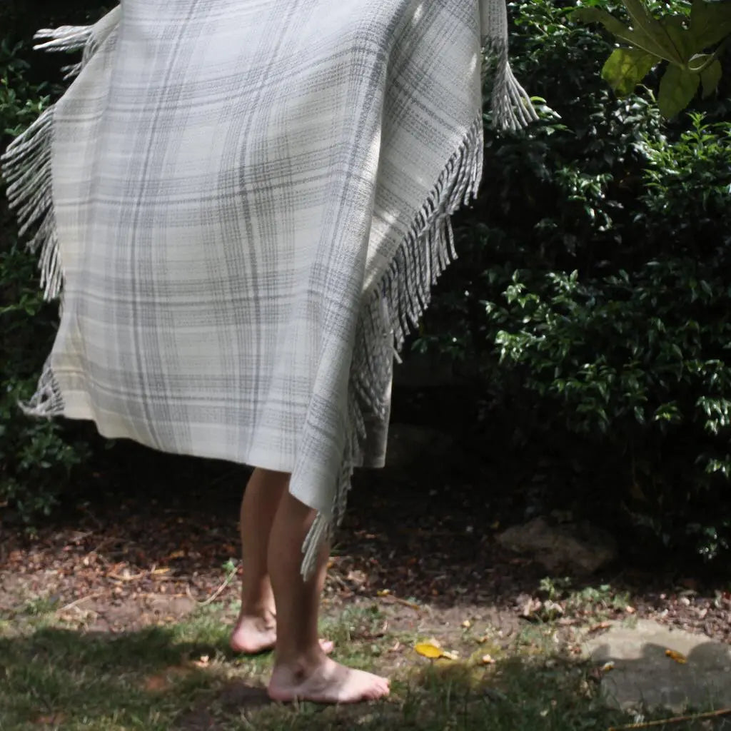 A person outdoors wearing a light gray and white plaid fringed wrap or poncho, standing barefoot on grass with bushes in the background.