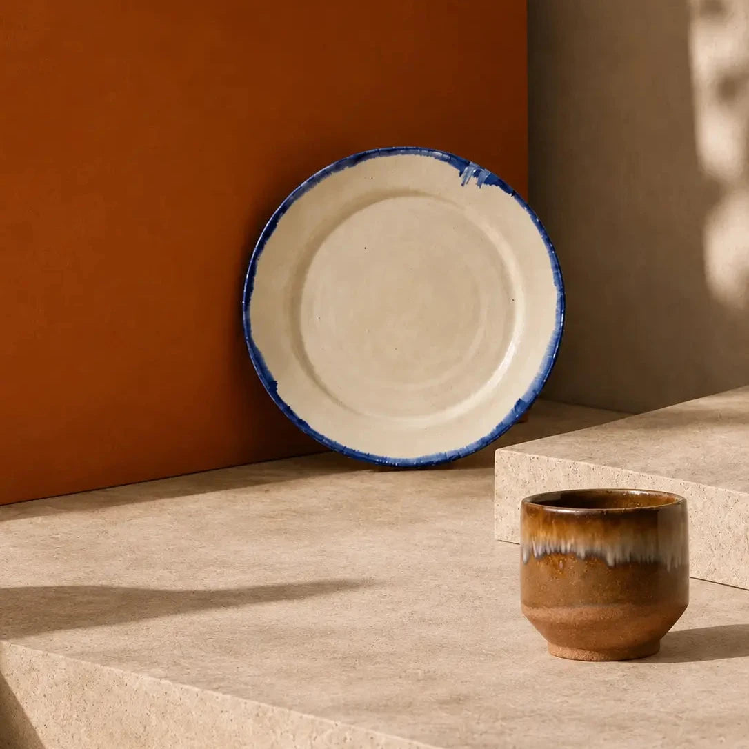 A styled collection of ceramic tableware, including mugs, a bowl, a plate, and a vase, arranged on beige stone steps against a warm terracotta and beige wall with leaf shadows. 