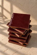 A stack of glossy square ceramic coasters in a deep burgundy red colour, arranged on a textured off-white surface.
