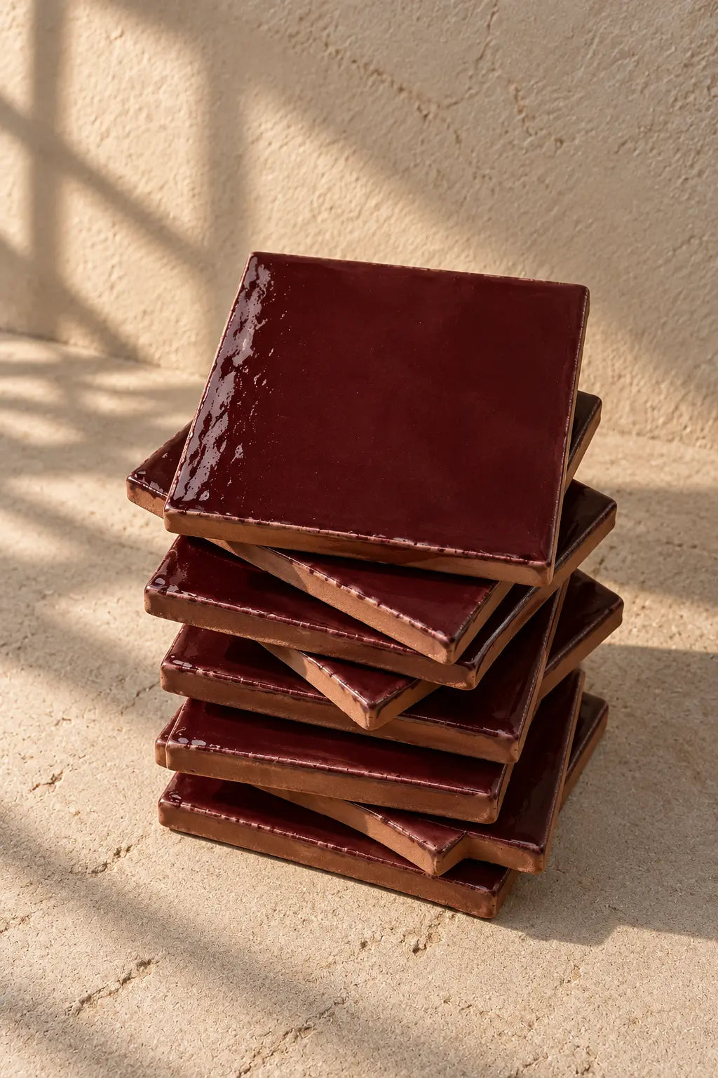 A stack of glossy square ceramic coasters in a deep burgundy red colour, arranged on a textured off-white surface.