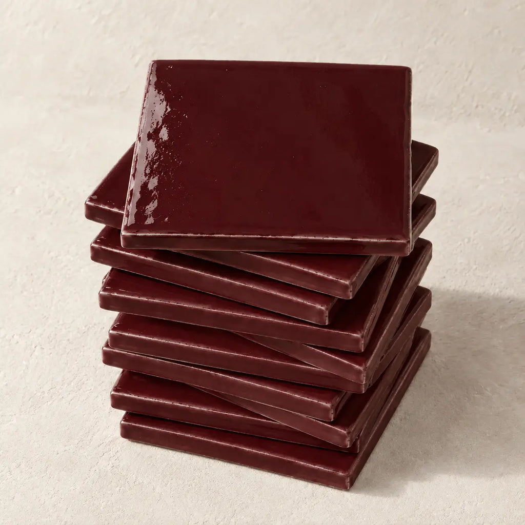 A stack of glossy square ceramic coasters in a deep burgundy red colour, arranged on a textured off-white surface.